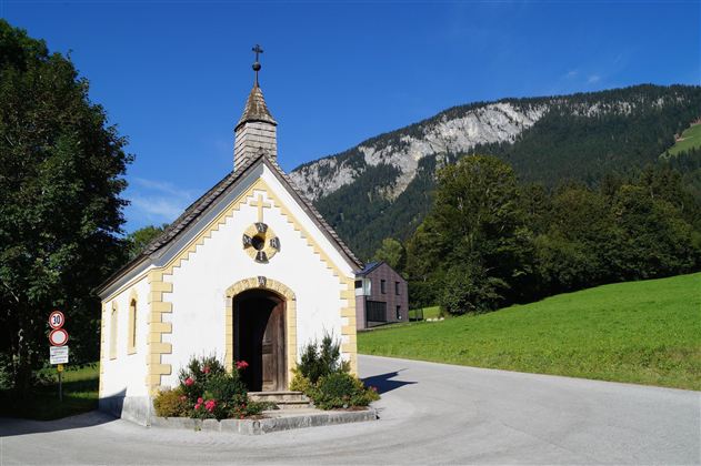 Eine kleine, charmante Kapelle steht an einer Straßenkreuzung inmitten von grünen Wiesen. Im Hintergrund erheben sich majestätische Berge unter einem klaren blauen Himmel.