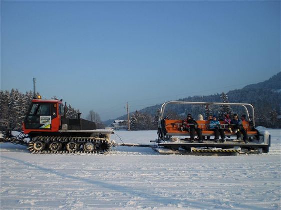 A snowmobile pulls a seating trailer with several people through a snowy landscape. The surroundings are wintry and clear, with mountains in the background.