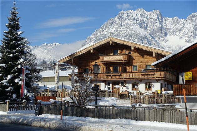 Ein schönes Holzhaus im Winter mit schneebedecktem Dach und malerischer Berglandschaft im Hintergrund. Der Himmel ist klar und blau.