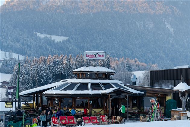 A cozy alpine hut in the snow with guests in front of the entrance. In the background, there are snow-covered mountains and trees visible.