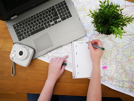 A desk with a laptop, a camera, and a folder. A hand is writing notes on a sheet of paper while a green plant stands in the background.