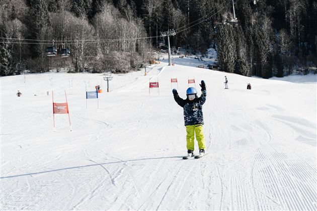 Dítě lyžuje na zasněžené sjezdovce a zvedá ruce do vzduchu. V pozadí jsou vidět další lyžaři a lyžařské svahy.