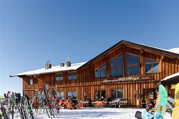 Eine gemütliche Berghütte aus Holz mit großen Fenstern und einer sonnigen Terrasse. Im Vordergrund stehen viele Skier, umgeben von schneebedecktem Gelände.