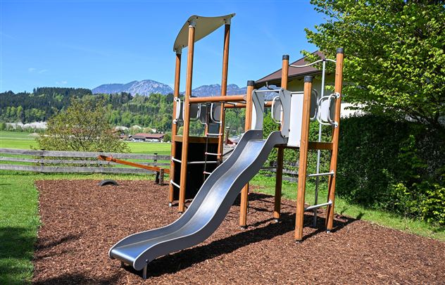 Een moderne speeltuin met een glijbaan van roestvrij staal en een houten frame. Op de achtergrond zijn bomen en bergen te zien.
