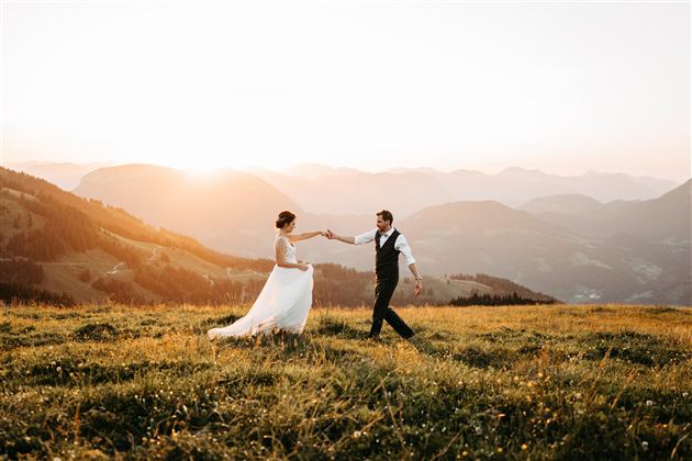 A bridal couple dances happily on a green meadow in the mountains. In the background, the setting sun glows.