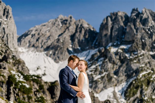 A bride and groom pose in front of an impressive mountain landscape. The peaks are snow-covered and the sky is clear.
