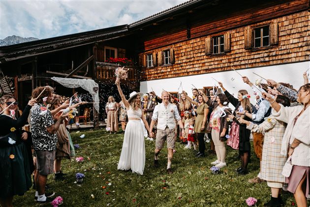 A bride and groom celebrate in front of a traditional wooden house. Surrounded by cheerful guests who cheer with flowers and confetti.