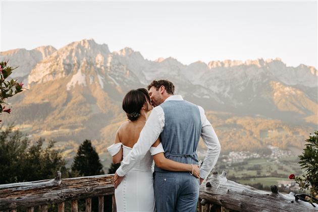 A bride and groom are standing on a porch and embracing each other. In the background, majestic mountains and a picturesque landscape can be seen.