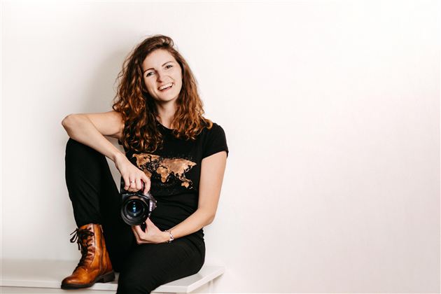 A smiling woman with curly hair is sitting on a table and holding a camera. She is wearing a black T-shirt and brown boots.