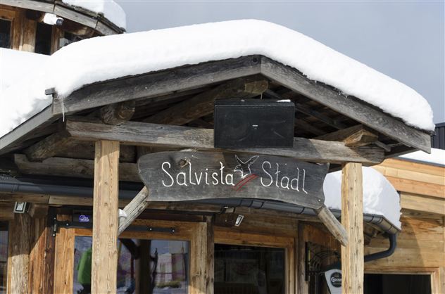 A wooden sign with the inscription "Salvista Stad" under a snow-covered roof. The surroundings depict a cozy, wintry atmosphere.