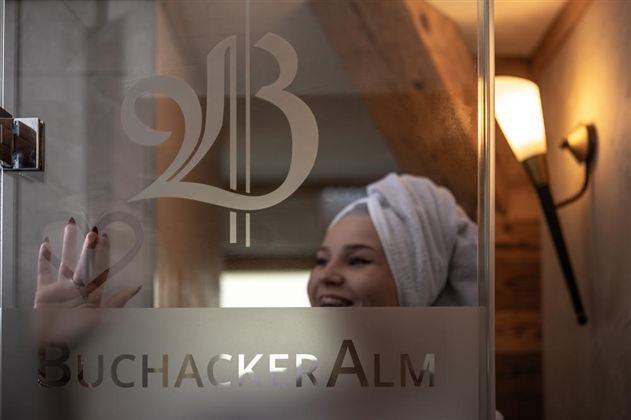 A woman with a towel on her head smiles behind a glass wall with the inscription "BuchackerAlm". Soft light illuminates the room, creating an inviting atmosphere.
