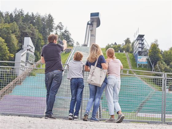 Eine Familie steht vor einer Sprungschanze im Freien. Im Hintergrund sind grüne Hügel und Sitzplätze zu sehen.