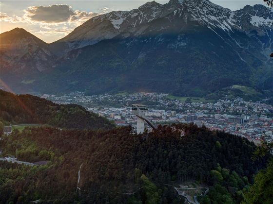 Eine malerische Aussicht auf eine Stadt umgeben von Bergen und Wald. Im Vordergrund ist eine Skisprungschanze zu sehen, die perfekt in die Landschaft integriert ist.