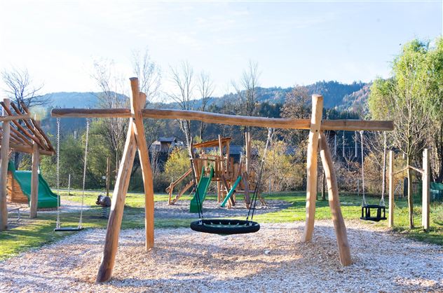 A playground with a wooden swing and various play equipment. In the background, there are trees and hills.