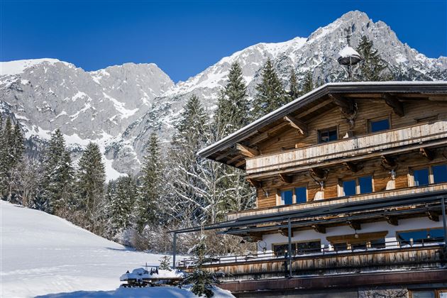 Ein charmantes Chalet im Schnee, umgeben von hohen Bergen und Nadelbäumen. Der Himmel ist klar und blau, was eine winterliche Atmosphäre schafft.