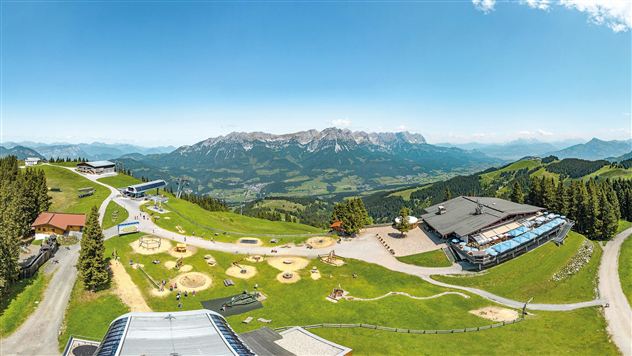 Een schilderachtig berglandschap met groene weilanden en met sneeuw bedekte toppen op de achtergrond. Op de voorgrond zijn er speeltuinen en gebouwen te zien die een prachtig uitzicht bieden.