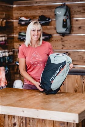 Zwei Personen stehen vor einem Geschäft mit Outdoor-Ausrüstung. Im Hintergrund sind Werbebanner und Verkaufsstände zu sehen.