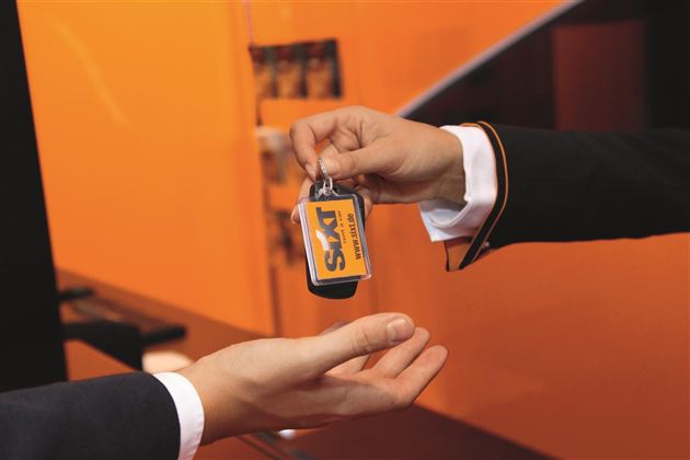 Eine Hand übergibt einen Schlüsselanhänger mit dem Logo von SKM. Der Hintergrund ist orange und schlicht.