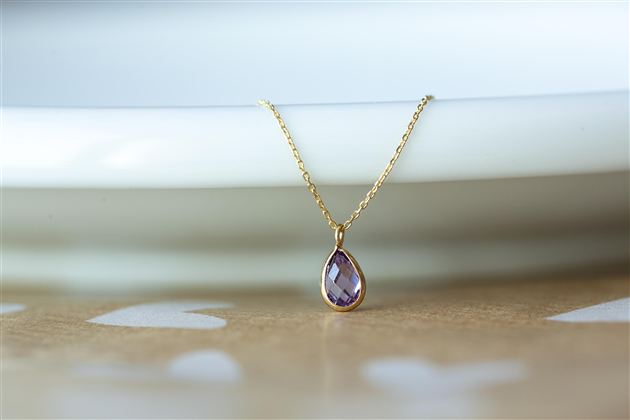 A delicate necklace with a teardrop-shaped amethyst pendant. The pendant is gold-colored and lies on a white plate.