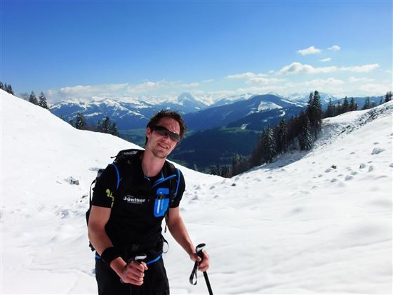 Ein Mann wandert auf einem schneebedeckten Berg mit Skistöcken. Im Hintergrund sind beeindruckende Berge und ein klarer Himmel zu sehen.