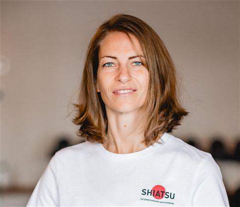 Eine Frau mit braunen Haaren steht lächelnd in einem hellen Raum. Sie trägt ein weißes T-Shirt mit dem Aufdruck "SHIATSU".