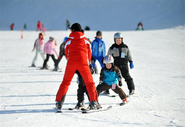 A ski school on a snowy slope with children in winter sports clothing. A teacher is helping the children with skiing.