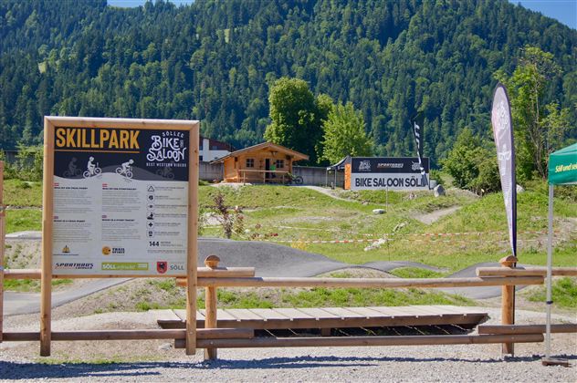 Ein Skillpark für Mountainbiker mit einem Informationsschild. Im Hintergrund sieht man die umliegenden Berge und einige Gebäude.