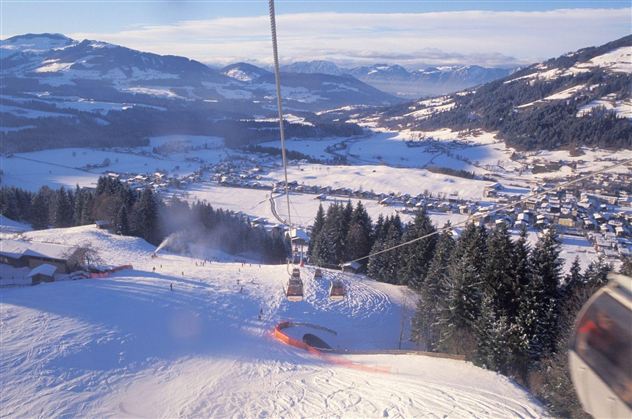 A picturesque snow landscape with a ski lift that rises above green fir trees. In the background, majestic mountains and a small town are visible.
