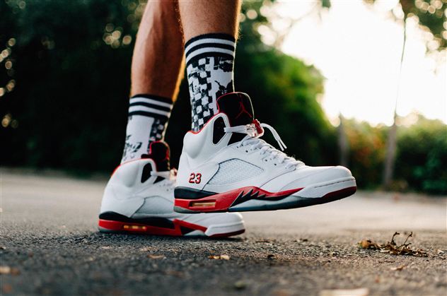 A pair of white sneakers with red accents is standing on an asphalt floor. The wearer is wearing colorful socks with a sporty pattern.