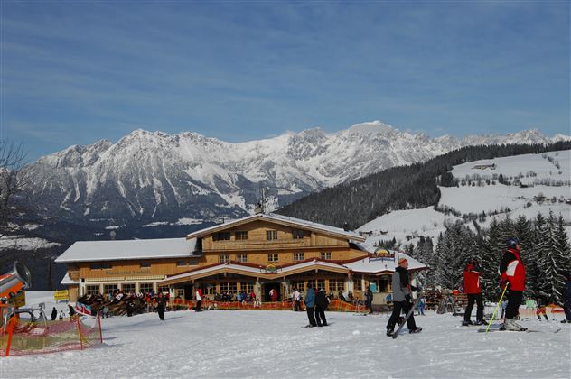 Eine alpine Skilandschaft mit einem gemütlichen Chalet und schneebedeckten Bergen im Hintergrund. Skifahrer und Besucher genießen das Winterwetter.