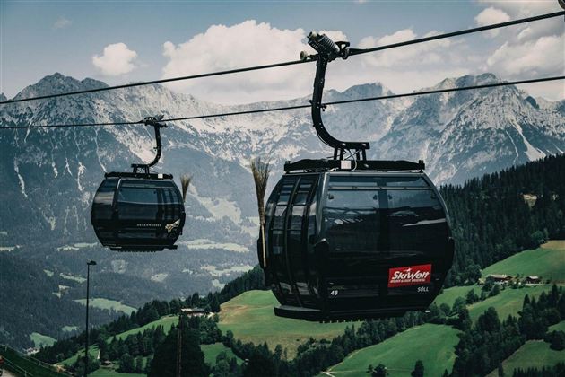 A cable car glides over green meadows and majestic mountains. In the background, tall, snow-capped peaks are visible.
