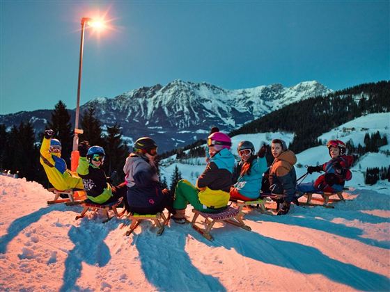 Een groep van zeven personen zit op sleetjes in de sneeuw. Op de achtergrond zijn bergen en een heldere avondlucht te zien.