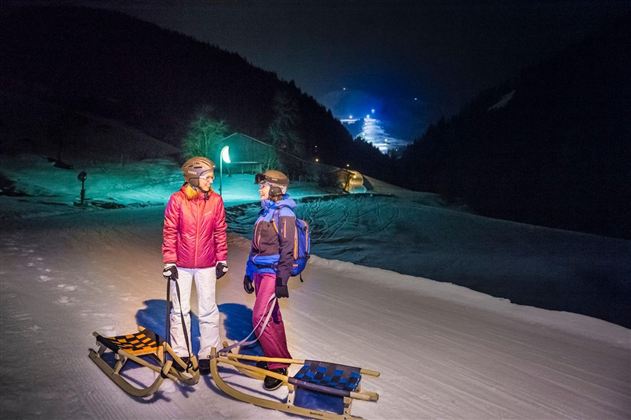 Twee personen staan 's nachts op een besneeuwde helling met sleetjes. Op de achtergrond schijnen lichten en een berglandschap is zichtbaar.
