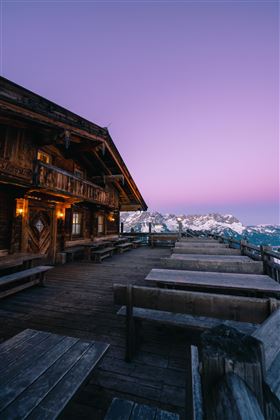 Een rustiek houten huis met een terras dat uitzicht biedt op de bergen. De lucht is gekleurd in zachte paarse en roze tinten.