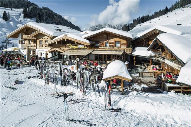 Een gezellige berghut in de sneeuw met veel ski's en wintersporters. Op de achtergrond zijn besneeuwde bergen en een heldere, blauwe lucht te zien.