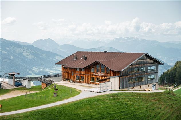 Ein großes Holzhaus auf einer grünen Anhöhe mit herrlichem Bergblick. Die Umgebung ist ruhig und natürlich, umgeben von sanften Hügeln und Bergen.