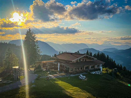 Eine charmante Berghütte umgeben von majestätischen Bergen. Die Sonne geht hinter den Gipfeln unter und taucht die Landschaft in warmes Licht.