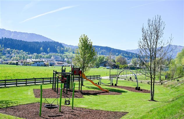 Een speeltuin met een glijbaan en schommels, omringd door groene weilanden. Op de achtergrond zijn bergen en een blauwe lucht te zien.