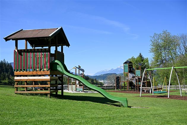 A playground with a wooden slide and swings outdoors. In the background, green meadows and trees can be seen.