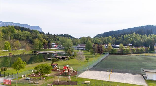 A quiet park with a playground and a large sports field. In the background, trees and mountains can be seen.