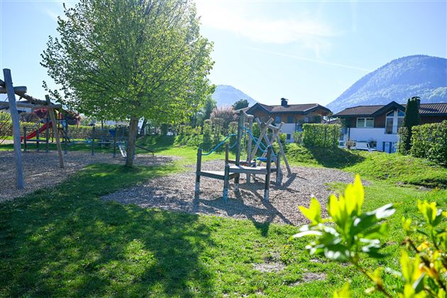 A playground in a green environment with a large tree. In the background, mountains and houses can be seen.