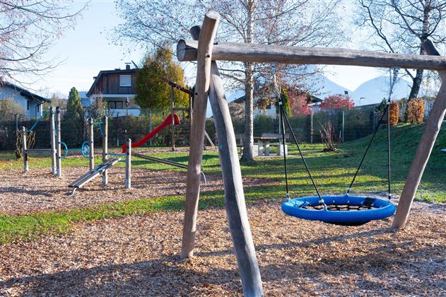 A playground with various play equipment, including a swing and a slide. The surroundings are green with trees and a quiet residential house in the background.