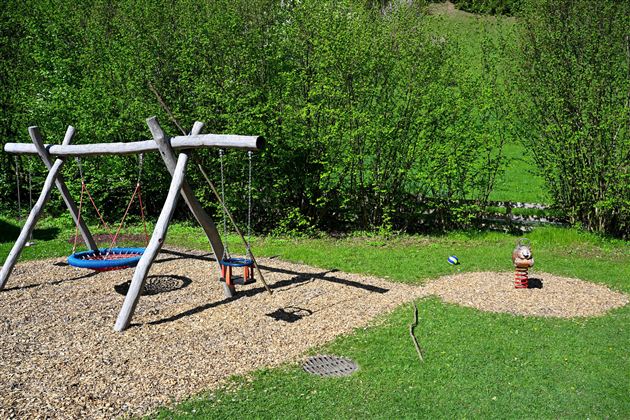 A playground with a wooden swing and a round sand area. In the background, green bushes can be seen.