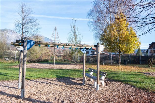A playground with a wooden climbing structure and a bench. In the background, there are trees and some buildings visible.