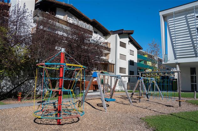 Ein Spielplatz mit einer Kletterstruktur und Schaukeln. Im Hintergrund sind mehrere Wohngebäude zu sehen.