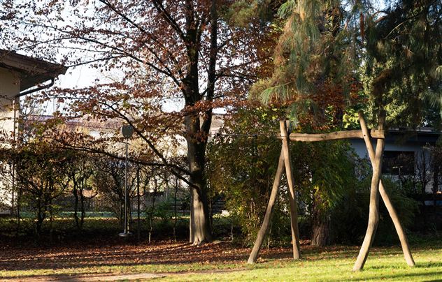 A playground with a wooden swing is located in a garden. In the background, trees and a house are visible.