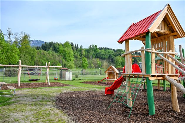 A beautiful playground with a wooden structure, slide, and swings. In the background, green meadows and trees can be seen.