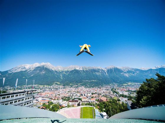 Ein Skispringer springt über eine malerische Stadt und die umliegenden Berge. Der Himmel ist klar und blau.