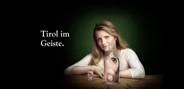 Een vrouw zit aan een tafel, achter een glazen fles met een gezicht. De achtergrond is donkergroen, en de tekst zegt "Tirol in Geest".