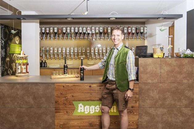 Een vriendelijke man in traditionele Beierse outfit staat achter de bar. Achter hem staan talrijke tapkranen voor bier opgesteld.